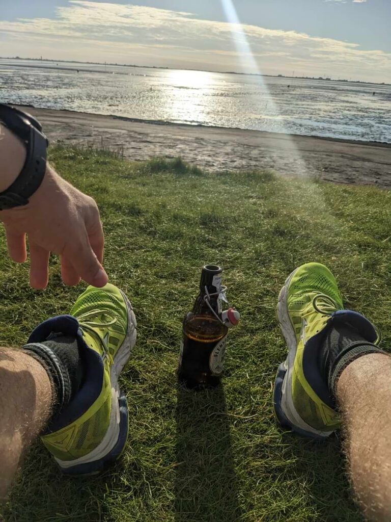 Mann sitzt au Wiese mit einem Bier zwischen den Füßen.